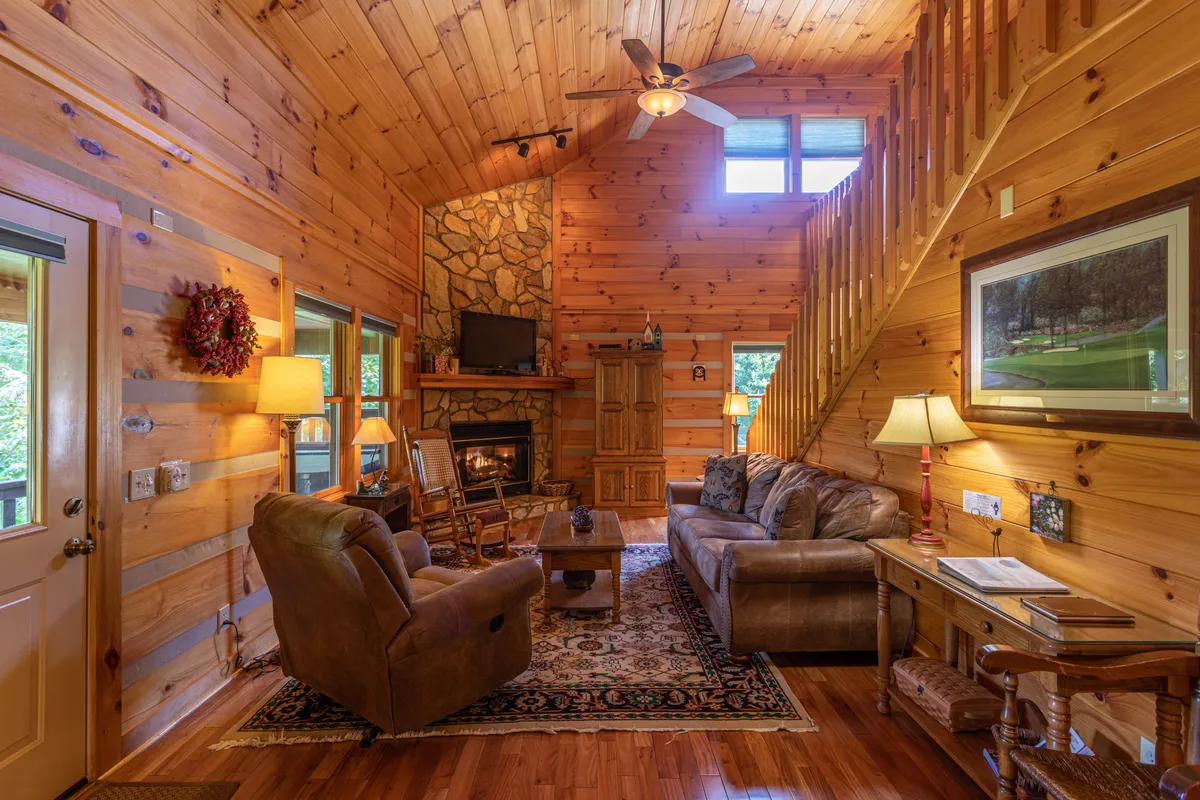 This image shows a warm, inviting living area with a flickering gas fireplace as its centerpiece, a comfortable sofa, and a wooden staircase leading to the upper level. Prominent large windows fill the space with natural light and offer stunning mountain views, reinforcing the connection to the Blue Ridge scenery. The overall mood is cozy and relaxed, perfect for gatherings and unwinding by the fire.