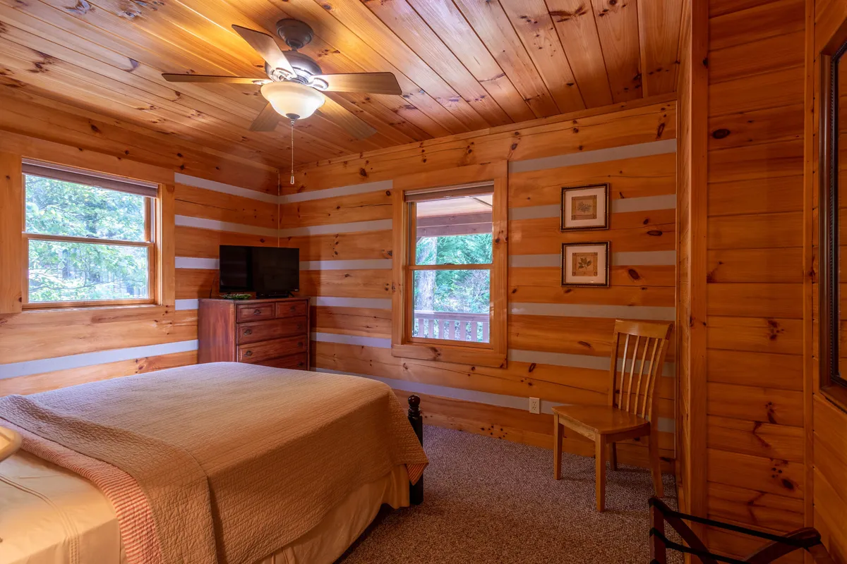 This cozy king bedroom features wooden walls, a large bed, sunlit windows, a dresser, and a chair, creating a warm and intimate atmosphere. The natural light streaming in emphasizes the peacefulness and comfort inside.
