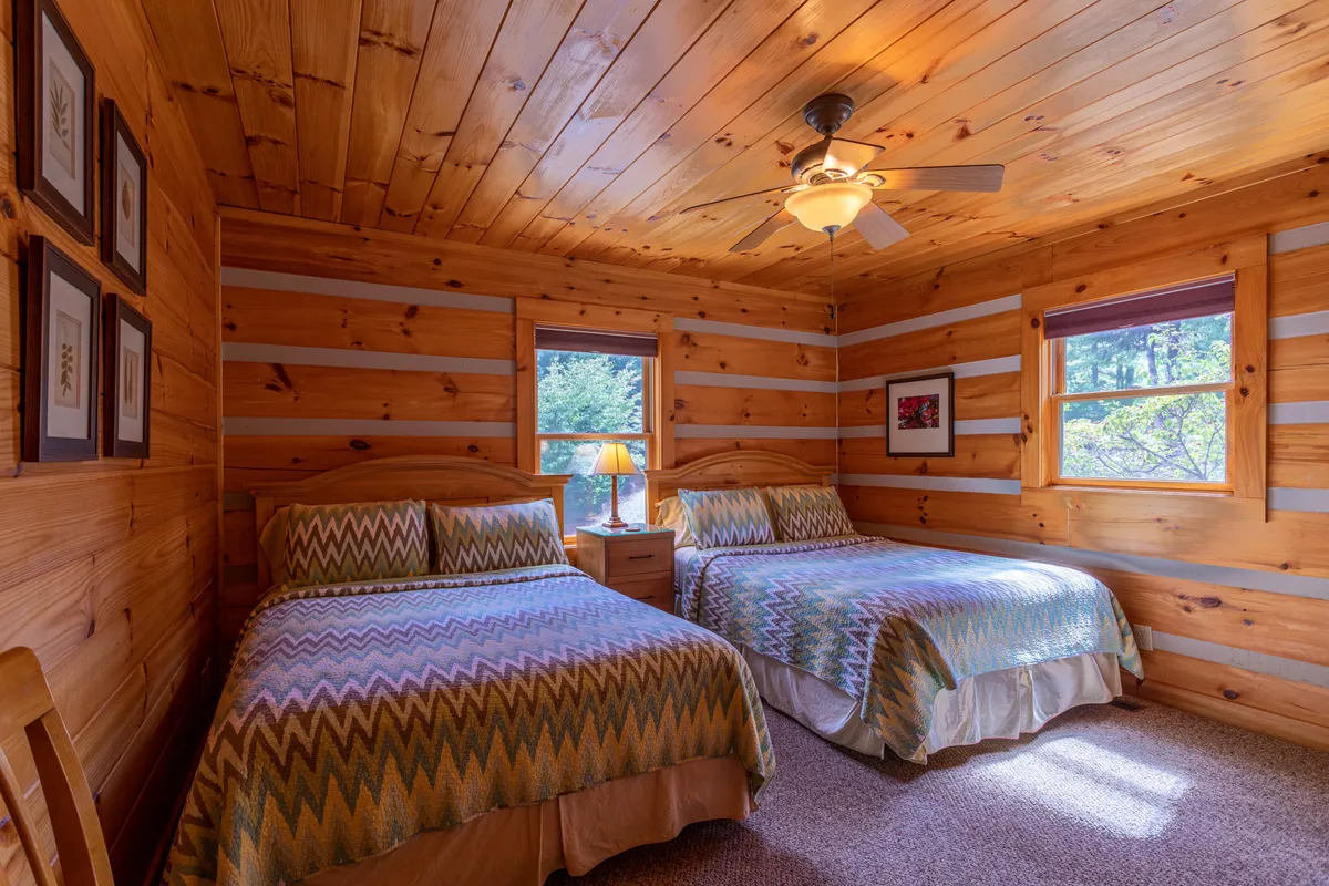 This bedroom features two beds with patterned comforters, wooden ceiling and walls, a lamp on a nightstand, and artwork on the wall. The setting feels comfortable and welcoming, with enough space for multiple guests or family members.