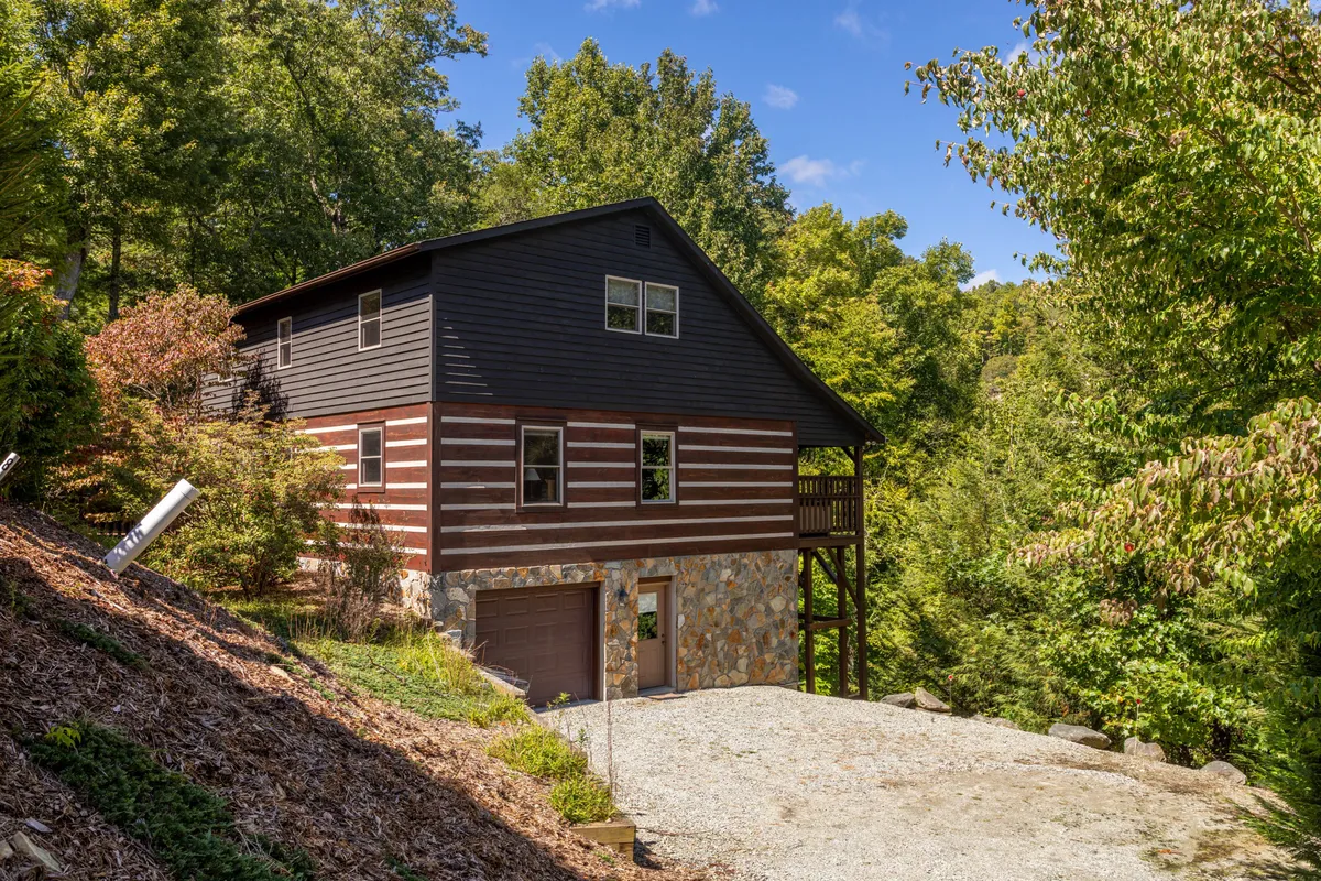 The image features the cabin's rustic log exterior, a stone foundation, and surrounding trees that immerse the property in natural beauty. The tranquil setting is emphasized by the abundance of greenery and the inviting structure.