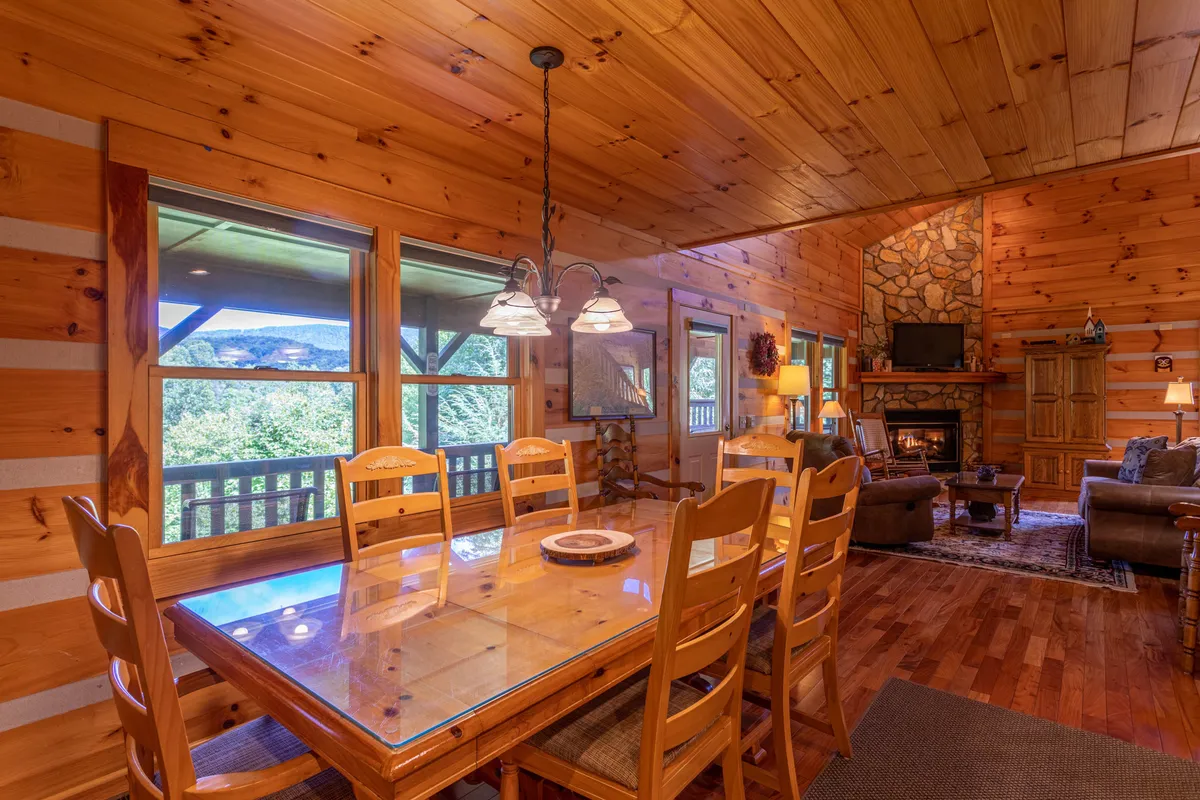 A beautifully styled dining table sits before a set of large windows, offering serene mountain vistas. A stone fireplace, wooden beams, and welcoming décor accentuate the warmth and comfort of the space, making it perfect for both intimate family dinners and larger celebrations.
