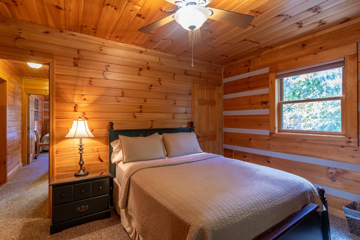 A serene bedroom highlighted by a plush bed, a glowing lamp, wooden walls, and a window that brings in natural light. The space exudes calm, inviting relaxation and restful sleep.