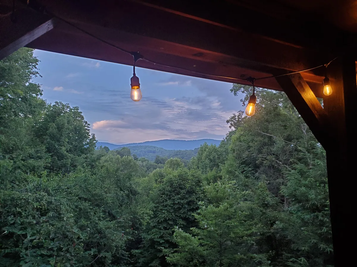 The hero image presents a tranquil outdoor dining area illuminated by warm, hanging light bulbs, set against the breathtaking backdrop of the Blue Ridge mountain range and lush greenery. The scene exudes serenity, with space for gathering, relaxing, and taking in the sunset, enticing guests to imagine cozy evenings or peaceful mornings surrounded by nature.