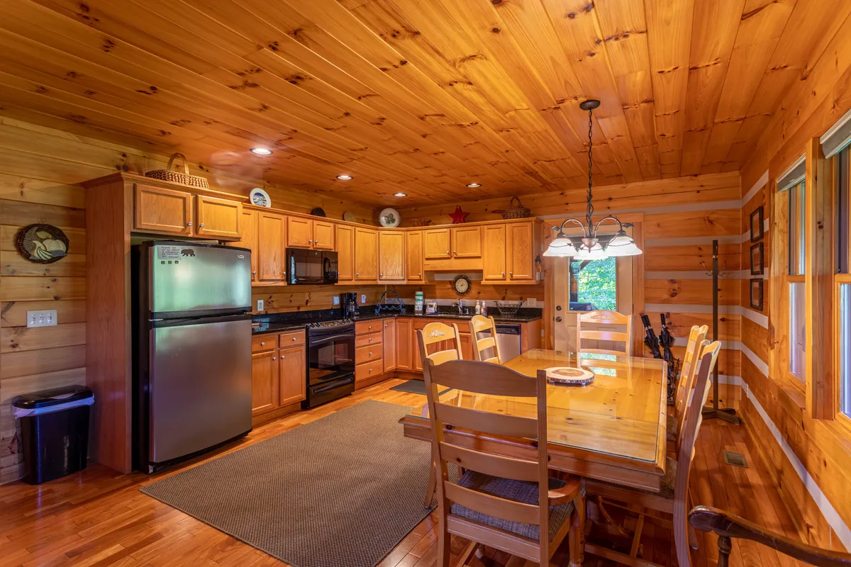 A spacious dining area centers around a grand wooden dining table surrounded by comfortable chairs, adjacent to an open-plan kitchen equipped with modern appliances and wooden cabinetry. This welcoming space is ideal for shared meals, family feasts, and game nights—the heart of the cabin's communal experience.